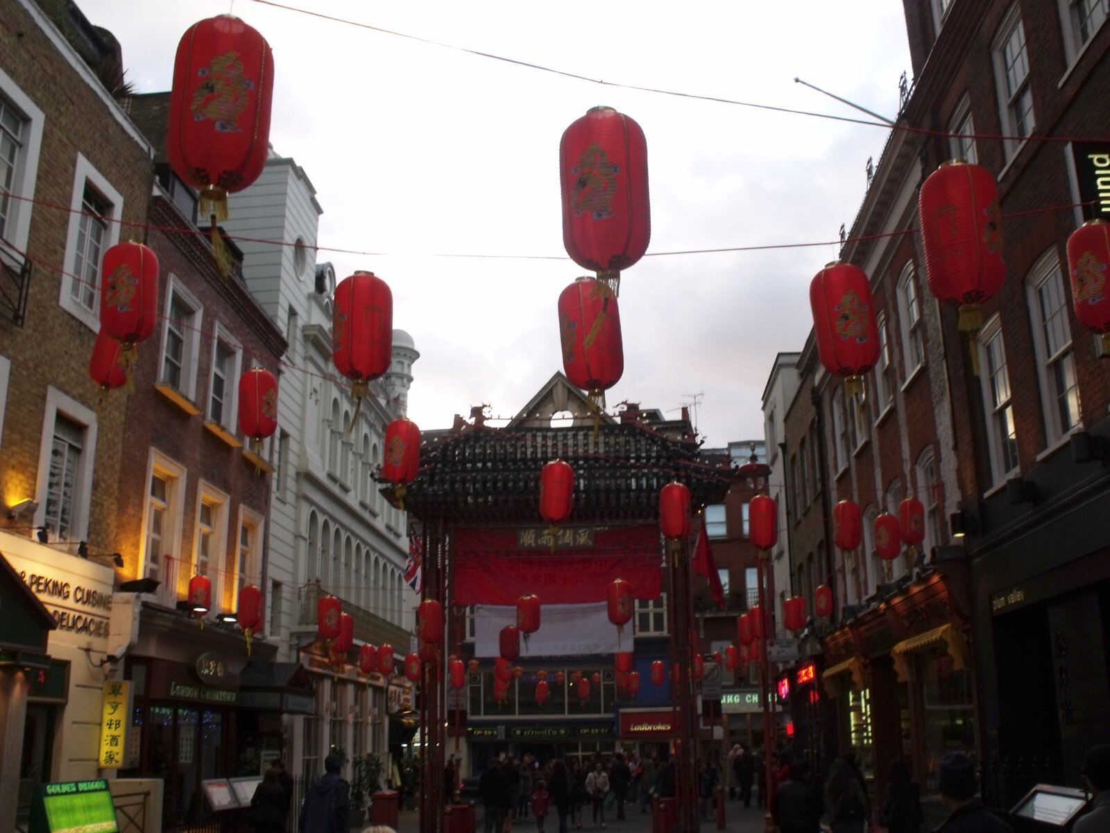 Red paper lanterns strung across Gerrard Street in London's Chinatown, with restaurant fronts on both sides and the Chinatown gate at the far end