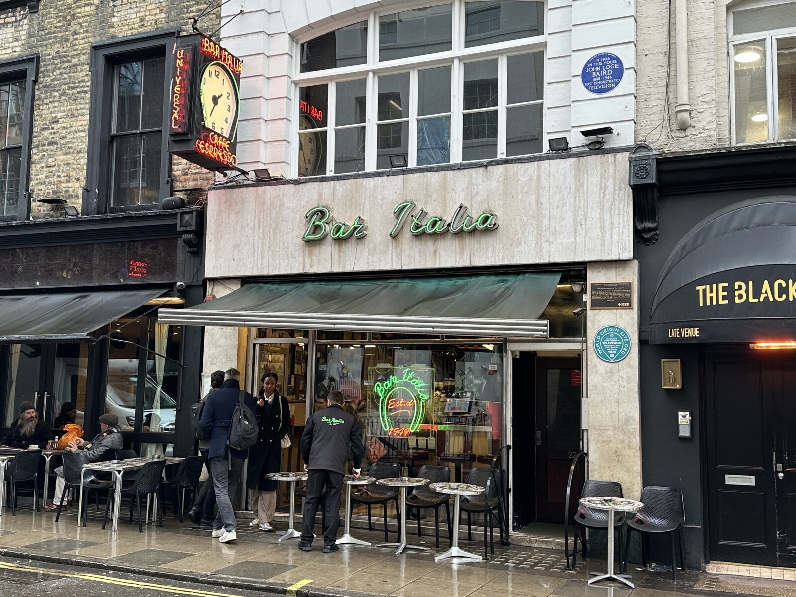 The front of Bar Italia on Frith Street in Soho, with its green neon sign, a large clock and pavement tables