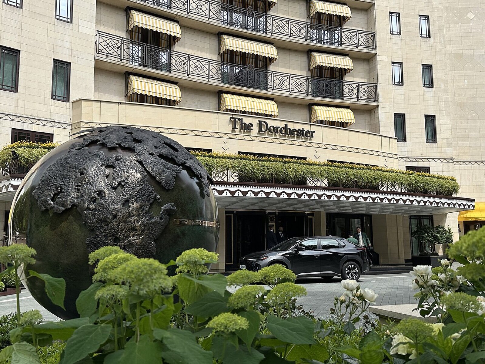 The cream facade of The Dorchester on Park Lane with striped awnings over the windows and a bronze globe sculpture in the garden in front