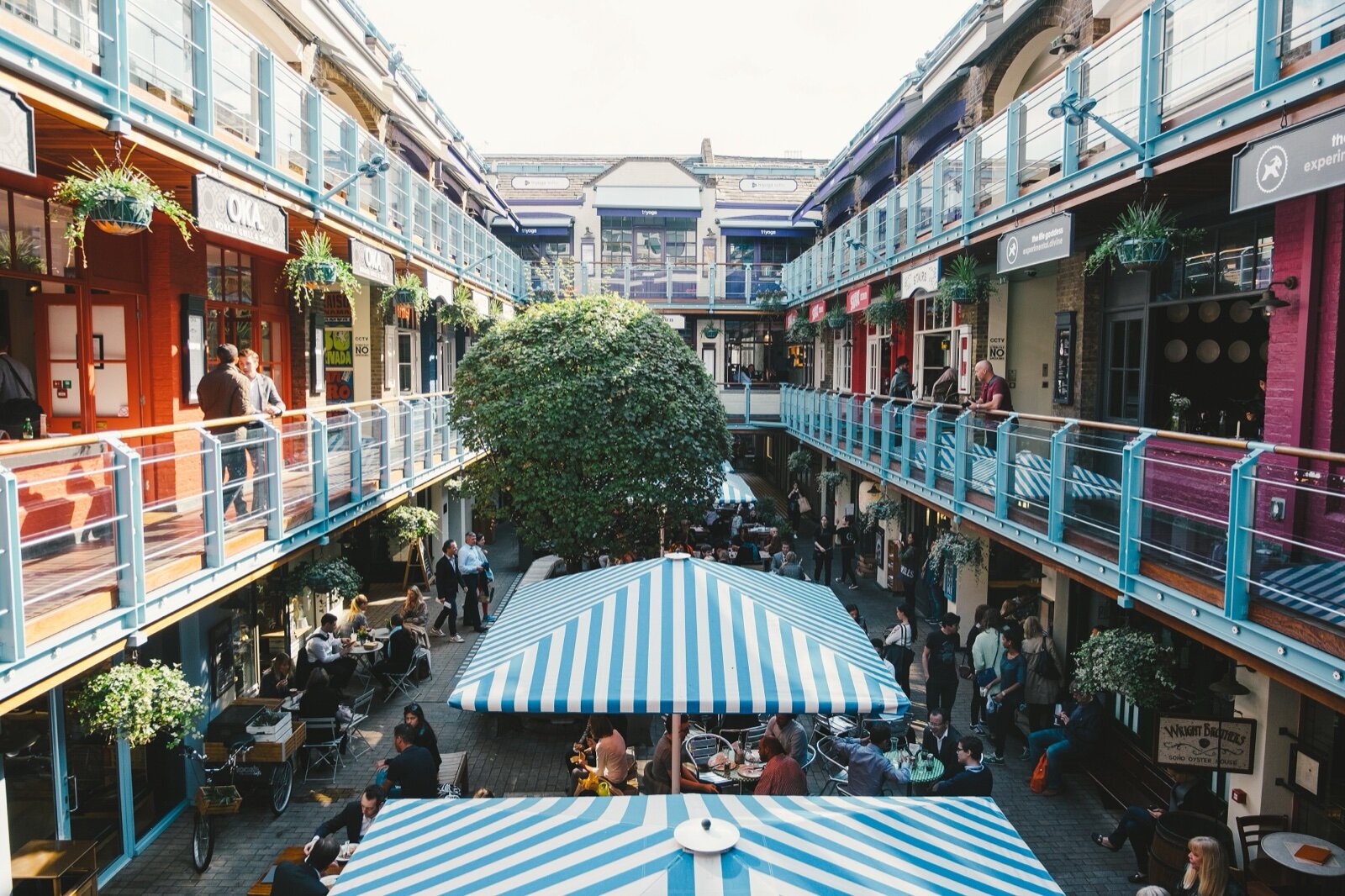 Kingly Court in Soho: a three-storey courtyard of restaurants with blue railings, hanging baskets, a round tree and striped parasols over outdoor tables