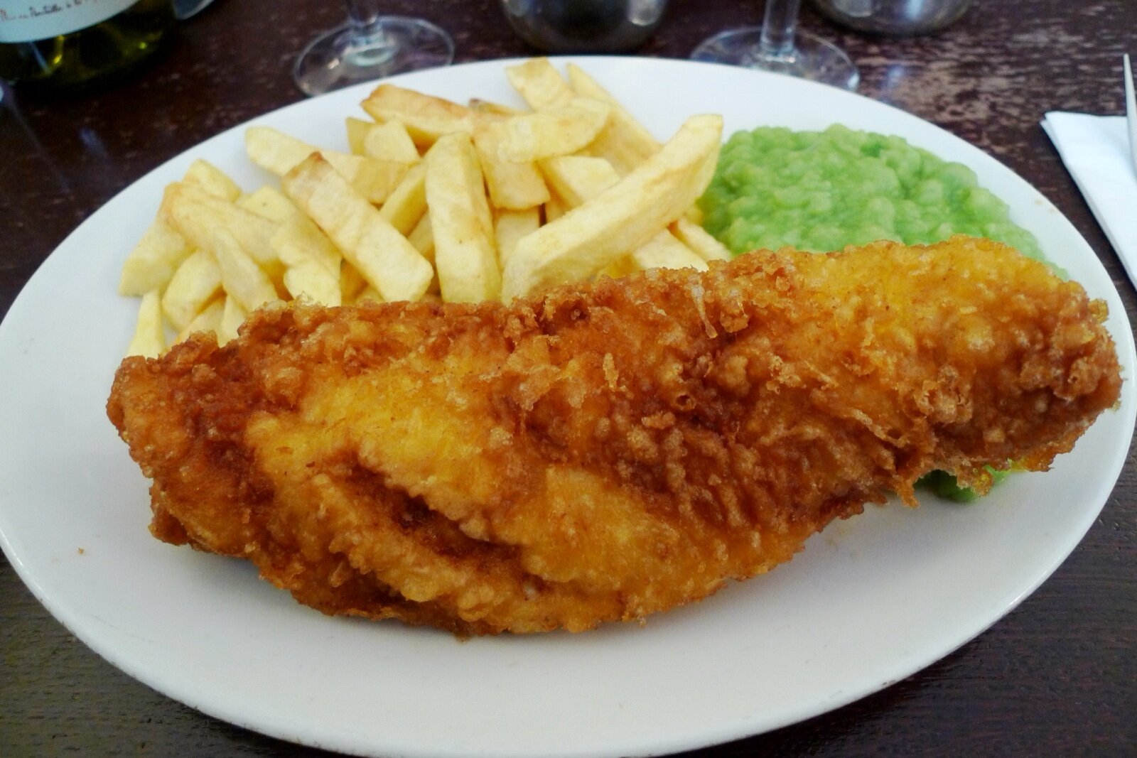 A large battered fish fillet with thick-cut chips and mushy peas on a white plate