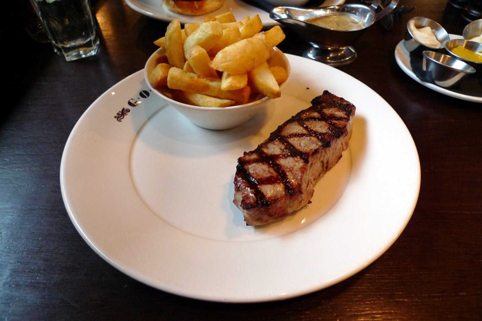 A chargrilled strip steak on a white plate beside a bowl of thick-cut chips, with sauce boats and mustard behind