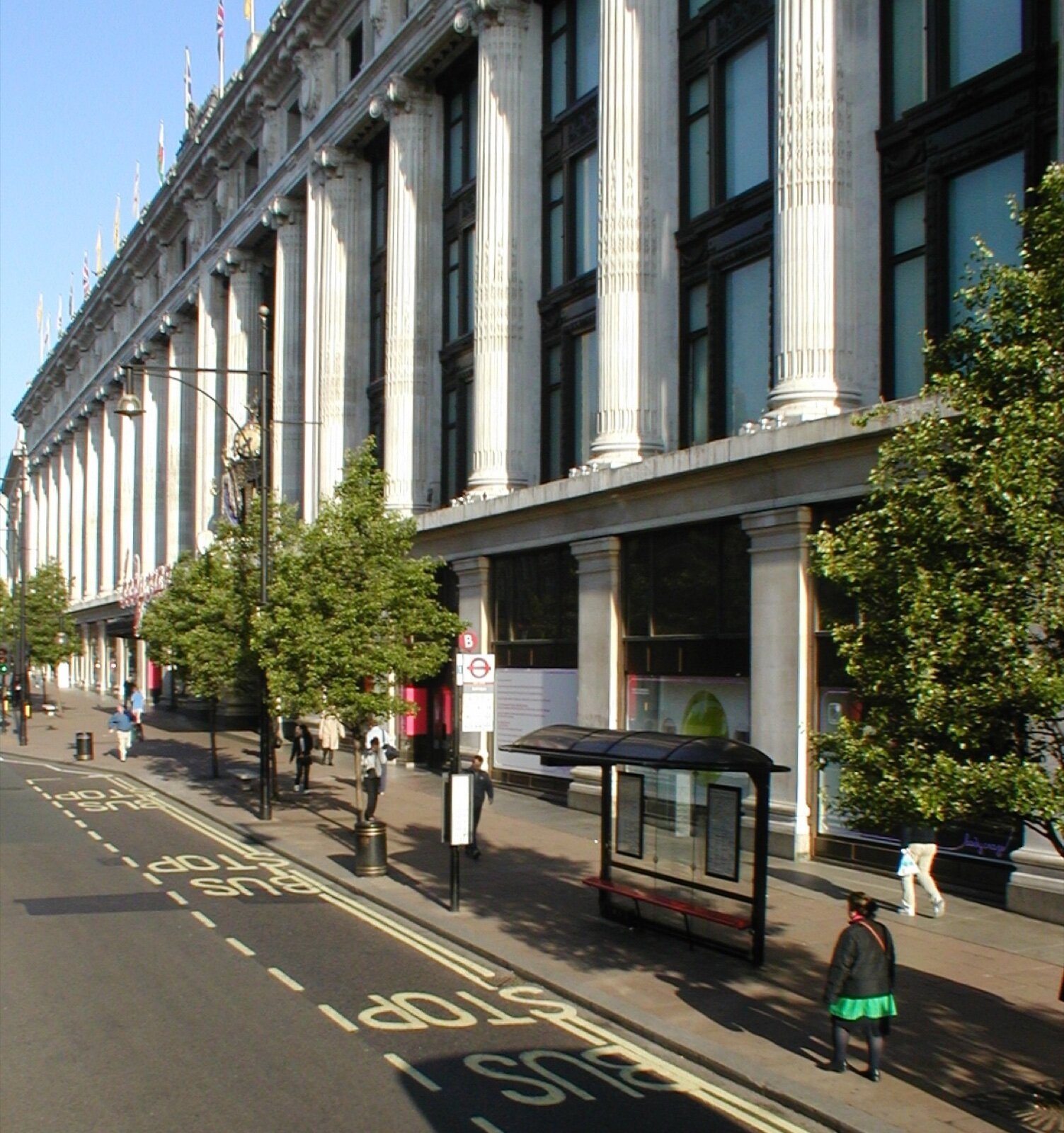 The long stone frontage of Selfridges on Oxford Street, with trees and a bus stop on the pavement