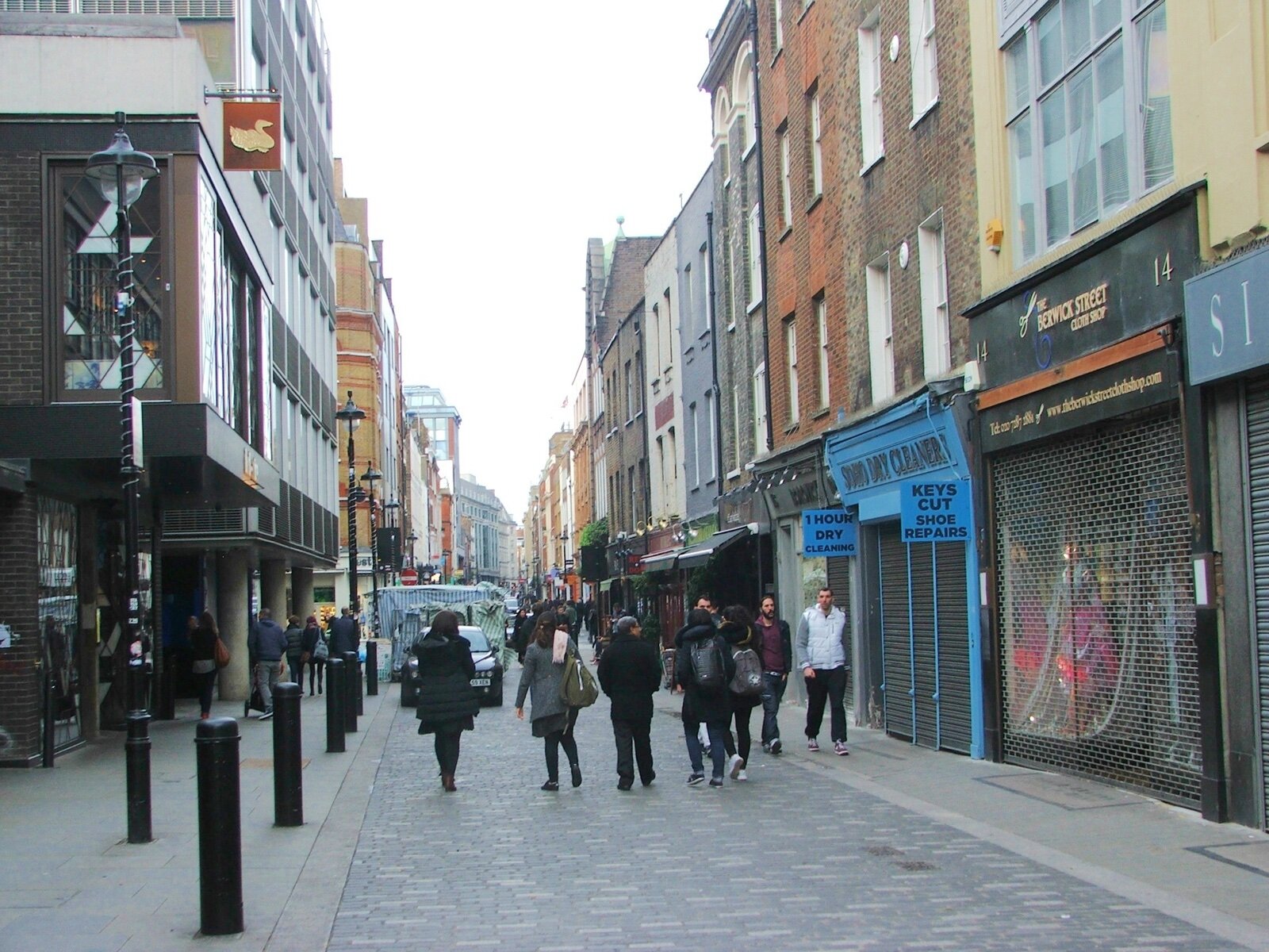 Berwick Street in Soho on a weekday, with market stalls, shuttered shopfronts and pedestrians on the paved street