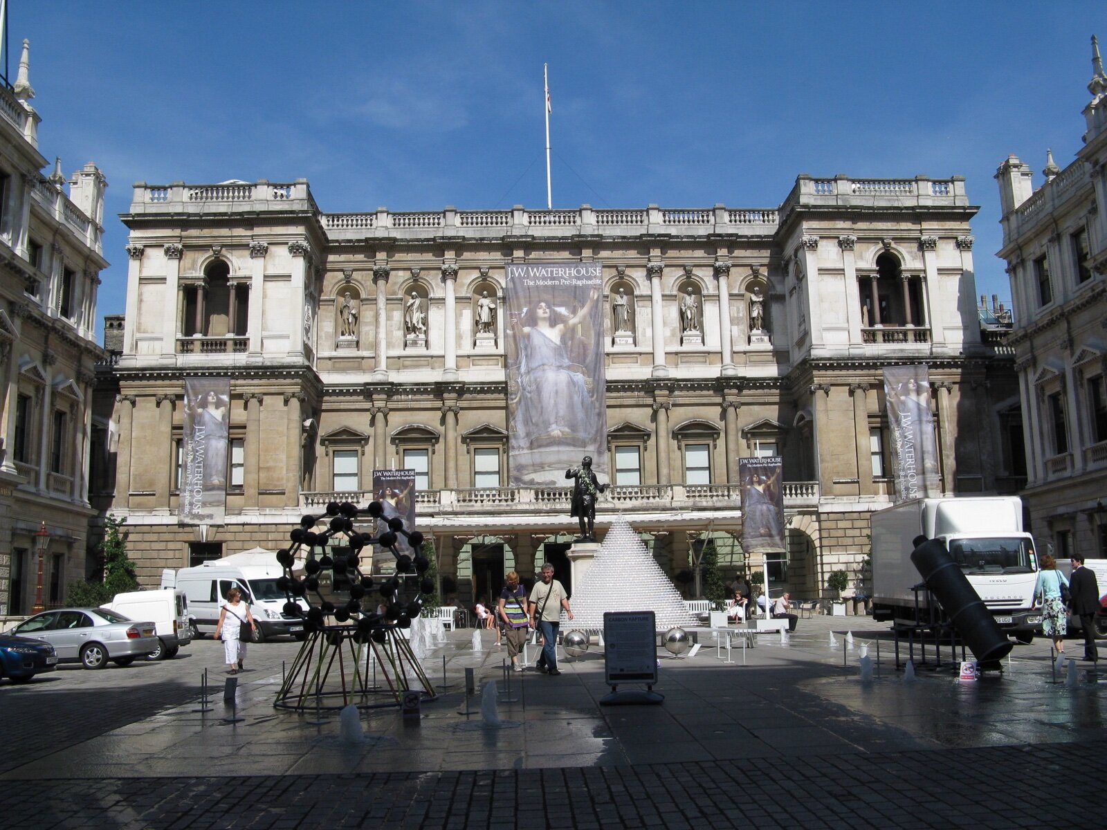 The stone facade of Burlington House on Piccadilly, home of the Royal Academy of Arts, seen across its courtyard on a sunny day with exhibition banners hanging from the front