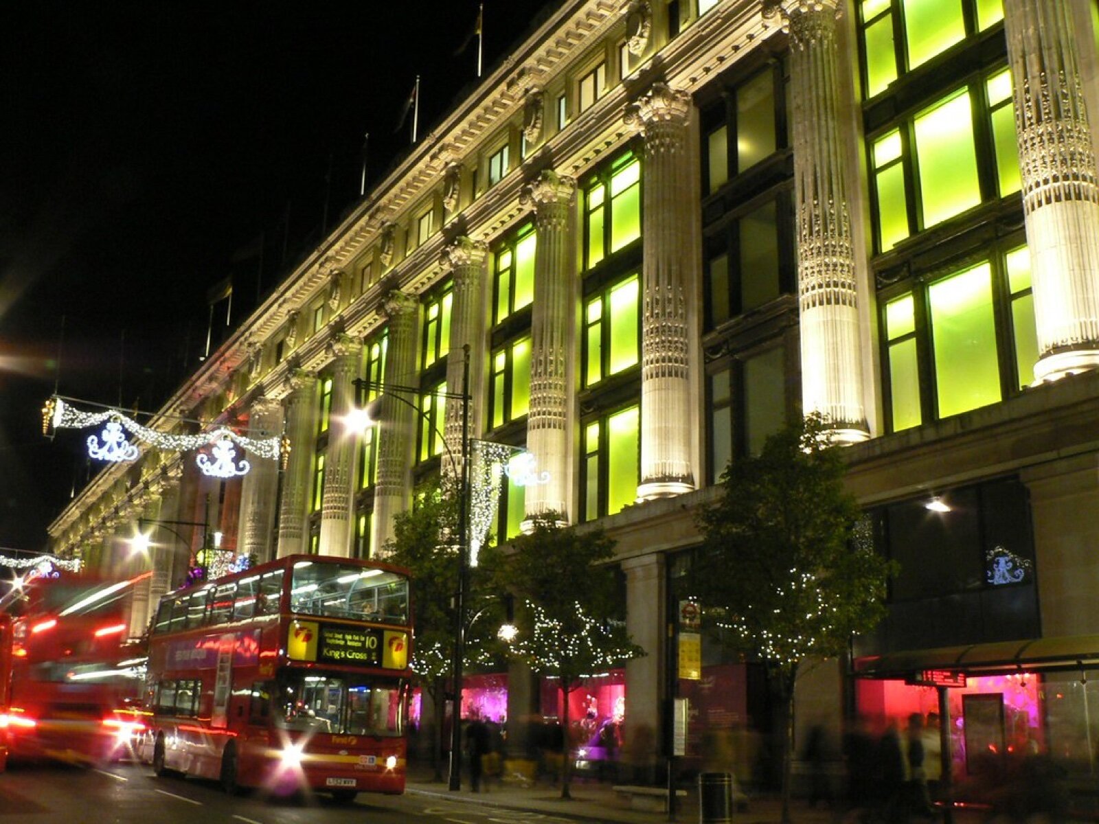 Selfridges on Oxford Street at night with a red double-decker bus passing and festive lights on the lamp posts